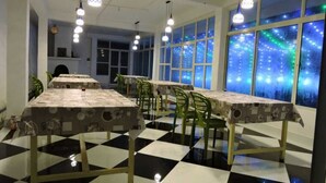 A dining area with checkered flooring, tables with patterned tablecloths, green chairs, and pendant lighting. - Vinayaka Inn Sittong (Kurseong)