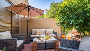 Un espace patio aménagé avec des meubles en osier, un canapé avec des coussins, un plateau avec des verres et un grand parasol.