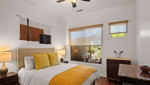 Une chambre à coucher comprenant un lit, des tables de chevet, un bureau, une chaise, un ventilateur de plafond et une fenêtre avec des stores.