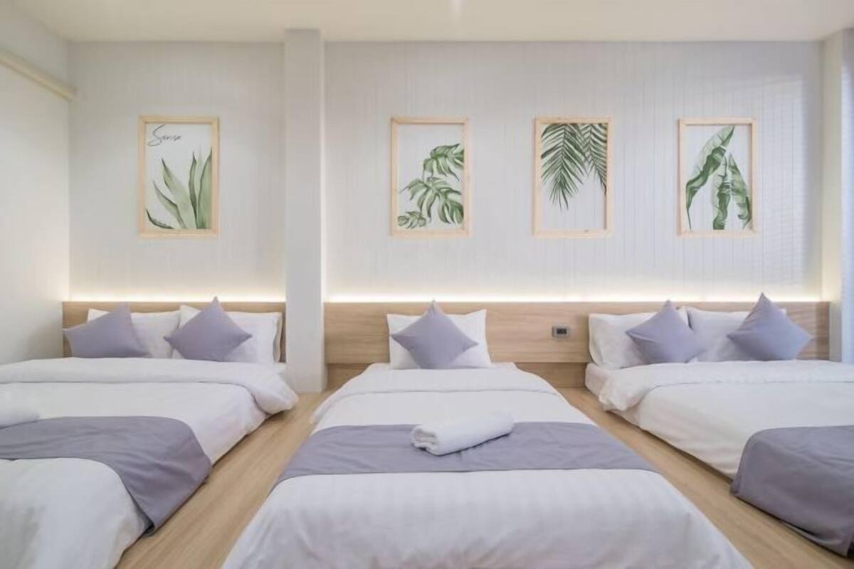 A hotel room with three beds, each with white bedding and purple pillows. The room features a wooden headboard, a wall-mounted light, and framed botanical artwork above the beds.