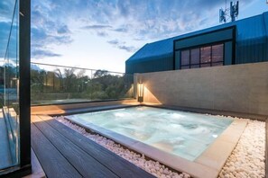 Un jacuzzi sur le toit, avec une terrasse en bois et des galets, entouré d’une balustrade en verre et d’un bâtiment moderne.