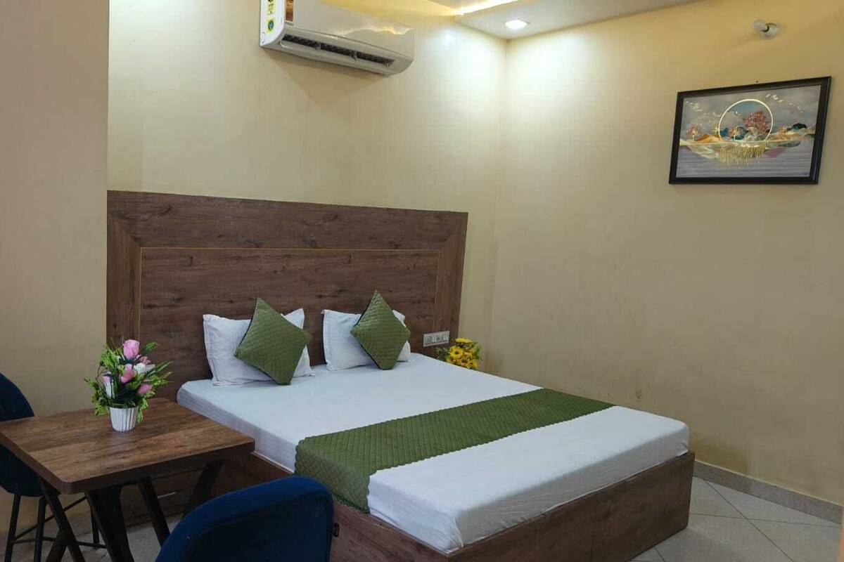 Een hotelkamer met een houten hoofdeinde, een bed met witte lakens en groene kussens, een bijzettafeltje met een bloevas, en een airconditioner aan de muur.