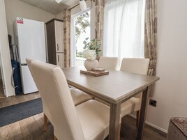 Un comedor con una mesa de madera, dos sillas blancas, un florero con flores y un refrigerador blanco al fondo.