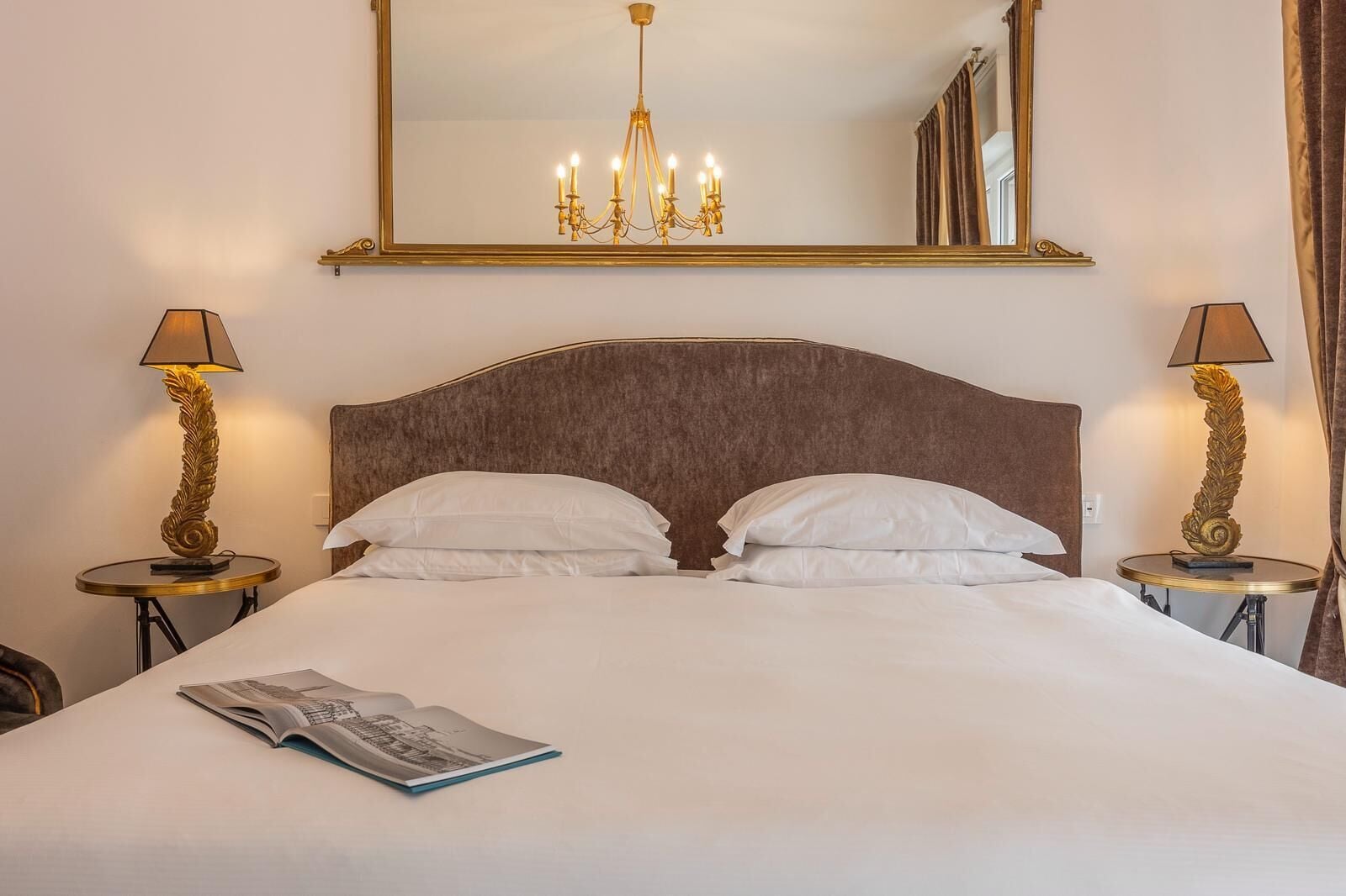 A hotel room with a large bed, two bedside tables with lamps, a chandelier, and a mirror on the wall.