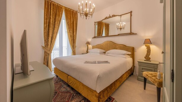 Habitació d'hotel amb un llit gran, una taula de treball amb un ordinador, una làmpada de cristall, un mirall i una taula petita amb una làmpada.