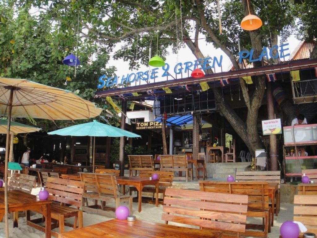 Un espace extérieur aménagé avec des tables et des chaises en bois, des parasols et une enseigne indiquant « SEA HORSE & GREEN PLACE ».