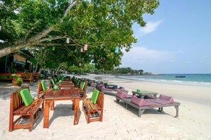 Ein Restaurant direkt am Strand mit Außenbereich, einschließlich Tischen und Stühlen, das unter einem großen Baum liegt.