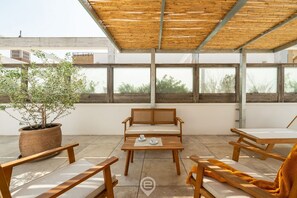 An outdoor patio area with wooden furniture, a potted plant, and large windows. - Stylish Terrace Apartment (Cagliari)