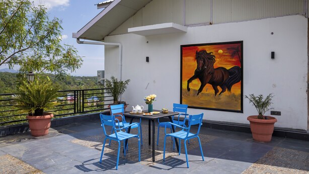A rooftop terrace with a table and chairs, potted plants, and a large painting of a horse. - Elivaas Esh Cottage Willow | Hillside With Lush Garden (Palampur)