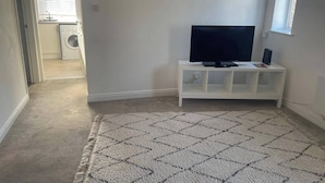 A room with a flat-screen TV on a white TV stand, a patterned rug, and a laundry area visible through an open door. - Horsforth Apartment Leeds Bradford Airport (Leeds)