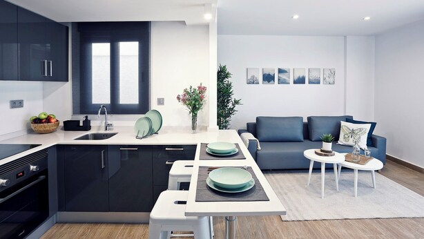 Une cuisine moderne avec des meubles de couleur sombre, un îlot central et un coin repas comprenant un canapé bleu et une table basse blanche.