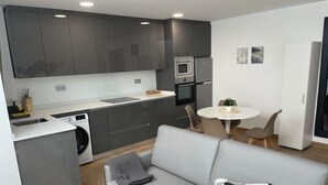 Une cuisine moderne avec des meubles gris, un four encastré et un four à micro-ondes. On y trouve un coin repas avec une table ronde et des chaises, ainsi qu’un canapé gris agrémenté d’un coussin à motifs.
