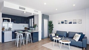 Une cuisine moderne avec des meubles de couleur sombre, un plan de travail blanc et un micro-ondes encastré. On y trouve un coin repas avec des tabourets blancs et une petite table blanche, sur laquelle sont posés des verres et une bouteille. Un canapé bleu et un tapis blanc se trouvent dans l’espace salon.
