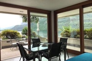 A dining area with a table and chairs, large sliding glass doors, and a view of a mountainous landscape. - Mergozzo Beach Bella Vista (Mergozzo)