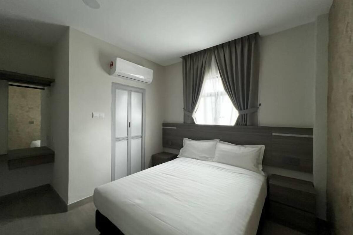 A hotel room with a bed, a nightstand, a window with curtains, an air conditioning unit, and a door.