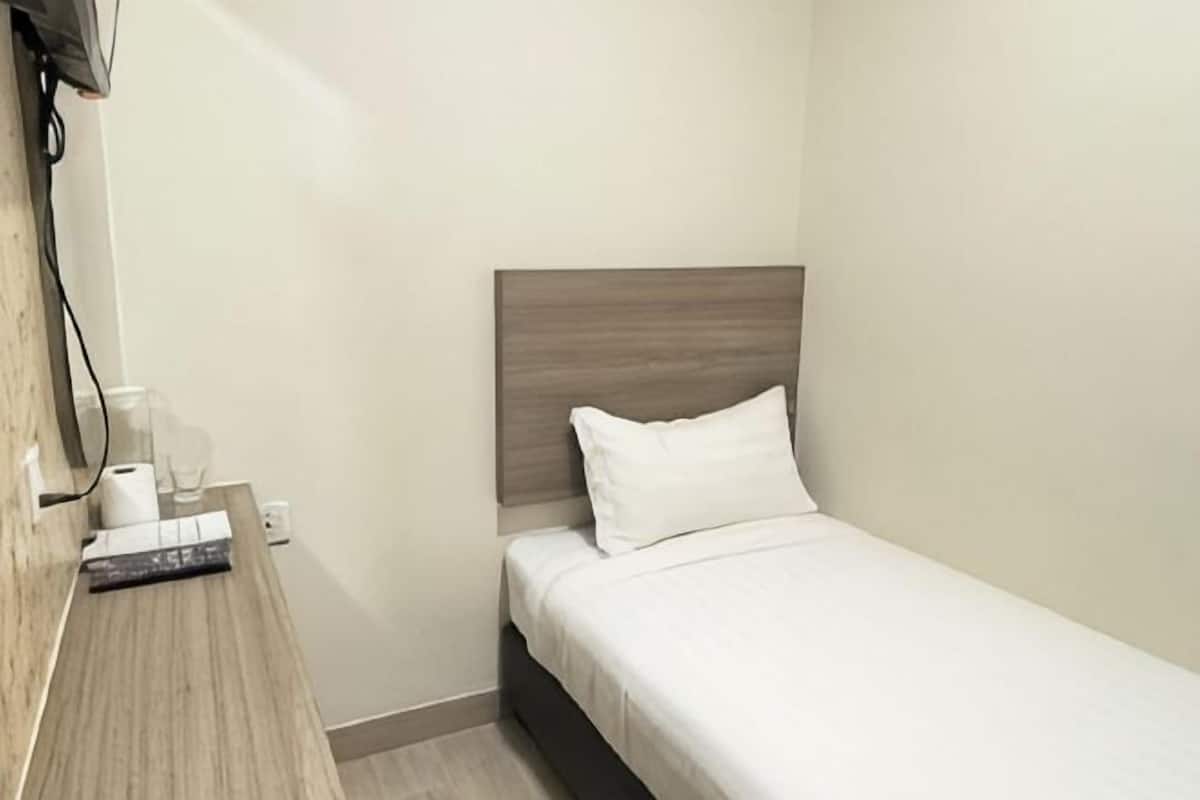 A small, single-bed room with a wooden headboard, a bedside table with a lamp, and a wall-mounted TV.