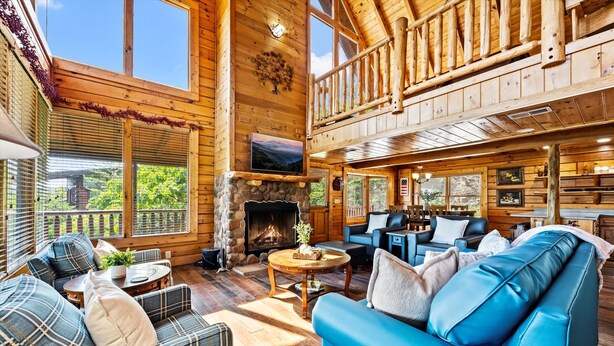 Interior espacioso de una cabaña de madera con chimenea, varias zonas de estar y grandes ventanas que ofrecen vistas del exterior.