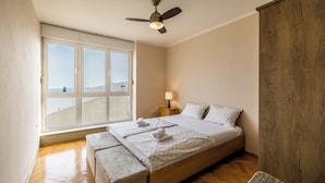 Une chambre avec une grande fenêtre, un lit avec des oreillers, une table de chevet avec une lampe et un ventilateur de plafond.
