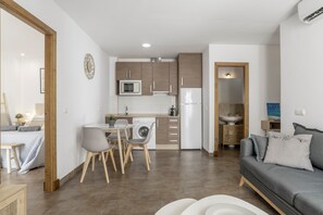 A modern kitchen with wooden cabinets, a microwave, and a refrigerator. There is a dining area with a table and chairs, and a living room with a sofa.