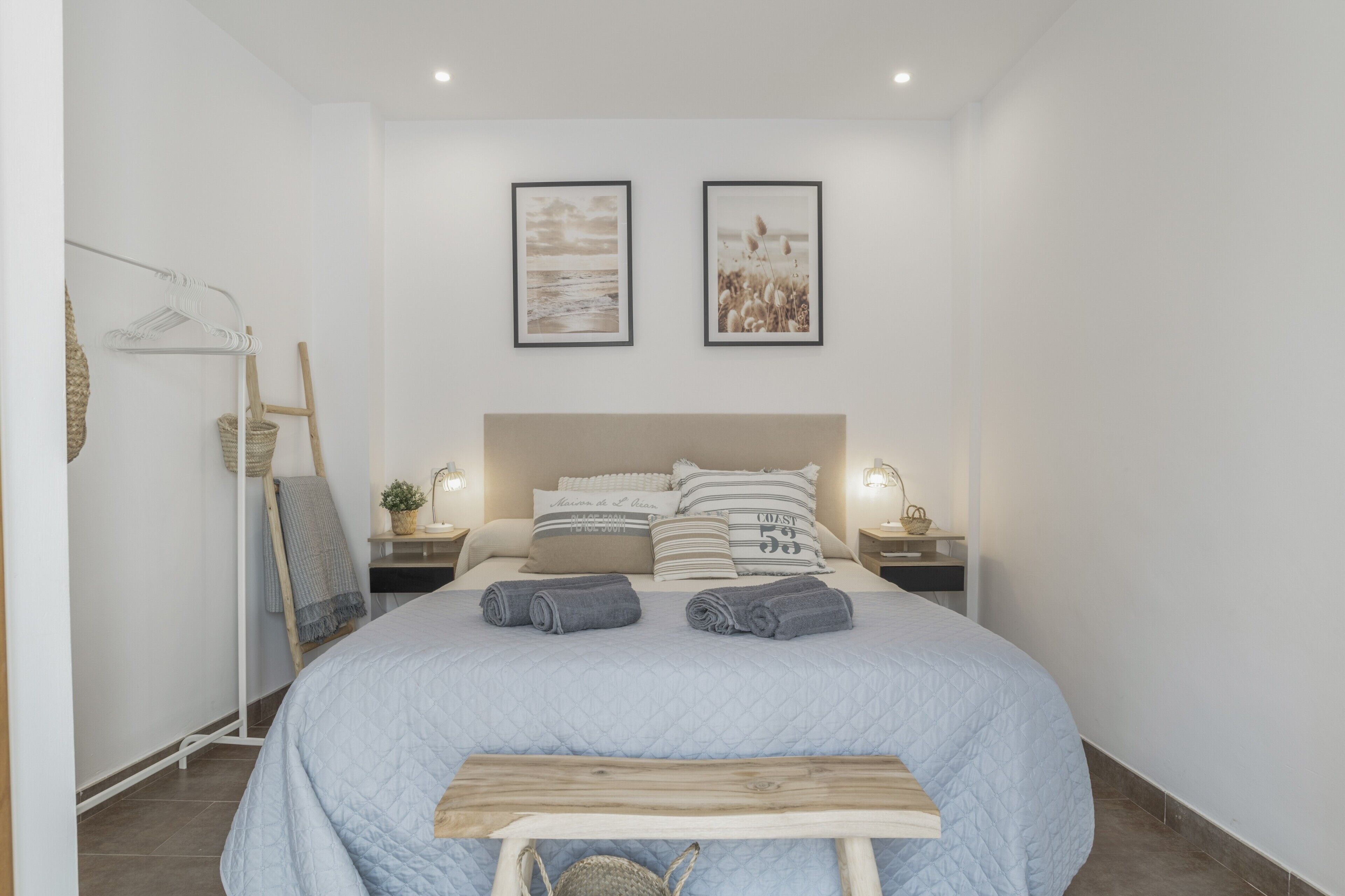 Ein Schlafzimmer mit einem Bett, zwei an der Wand befestigten Bildern, einem Nachttisch mit Lampe und einer hölzernen Bank.