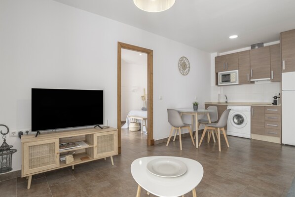 A modern living space with a flat-screen TV, dining table, chairs, a washing machine, and a kitchen area.