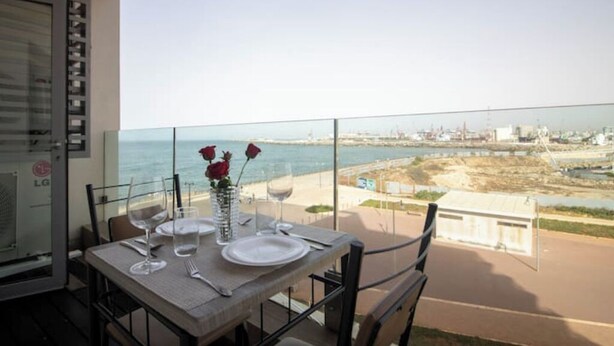Mesa de balcón dispuesta con platos, vasos y un florero de rosas rojas, con vistas a una zona costera con edificios y una playa.