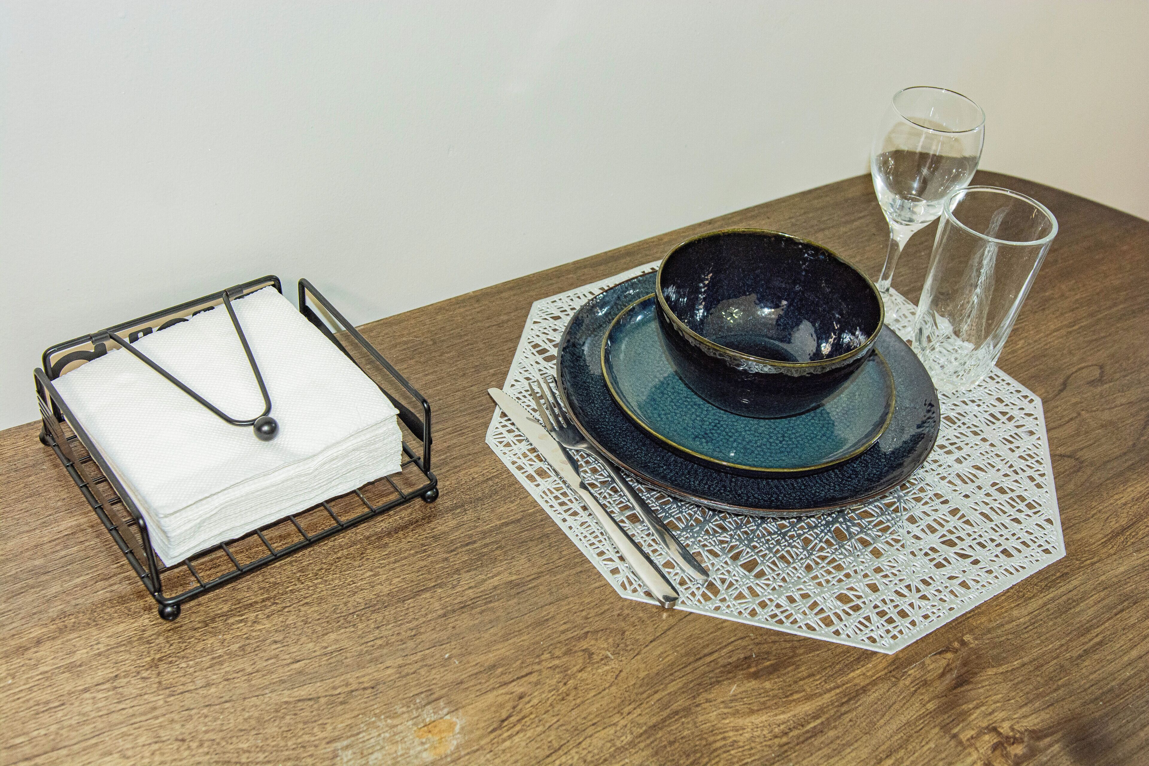 Une table à manger bien dressée, avec un bol bleu, deux verres à vin, un porte-serviettes et une nappe en dentelle.