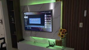 Een woonkamer met een flatscreen televisie aan een witte muur, een decoratieve vlinder, een bloemistisch kunstwerk aan de muur en een vaas met zonnebloemen op een plank onder de televisie.