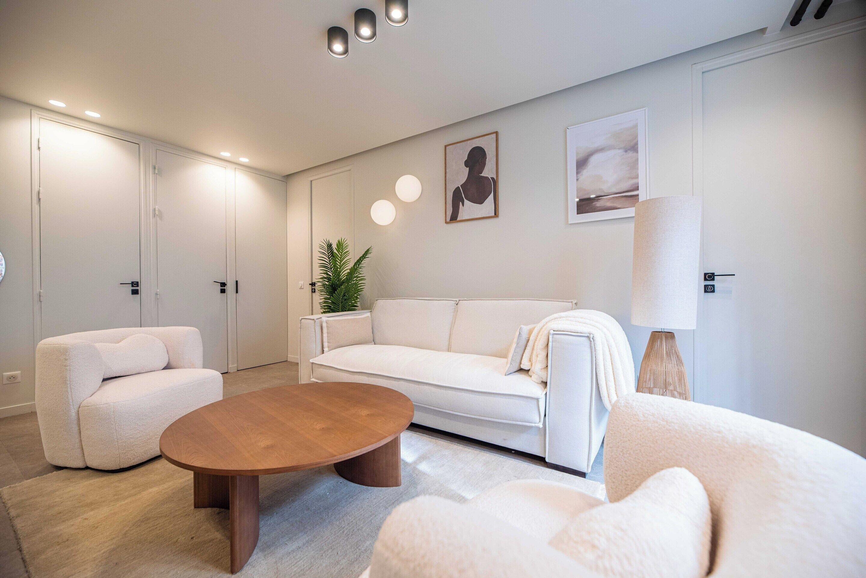 Ein modernes Wohnzimmer mit einem weißen Sofa, einem runden Holztisch und einem weißen Sessel. Im Hintergrund sind zwei Türen, eine Zimmerpflanze und Wandbilder zu sehen.