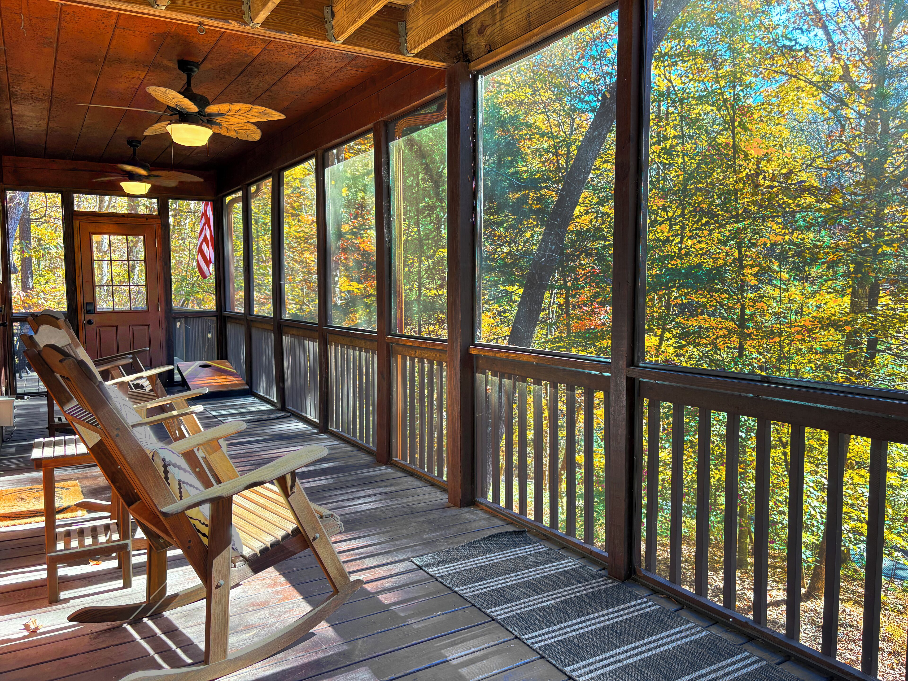 Une terrasse couverte avec des chaises en bois et une table, de grandes fenêtres offrant une vue sur les arbres aux couleurs automnales.