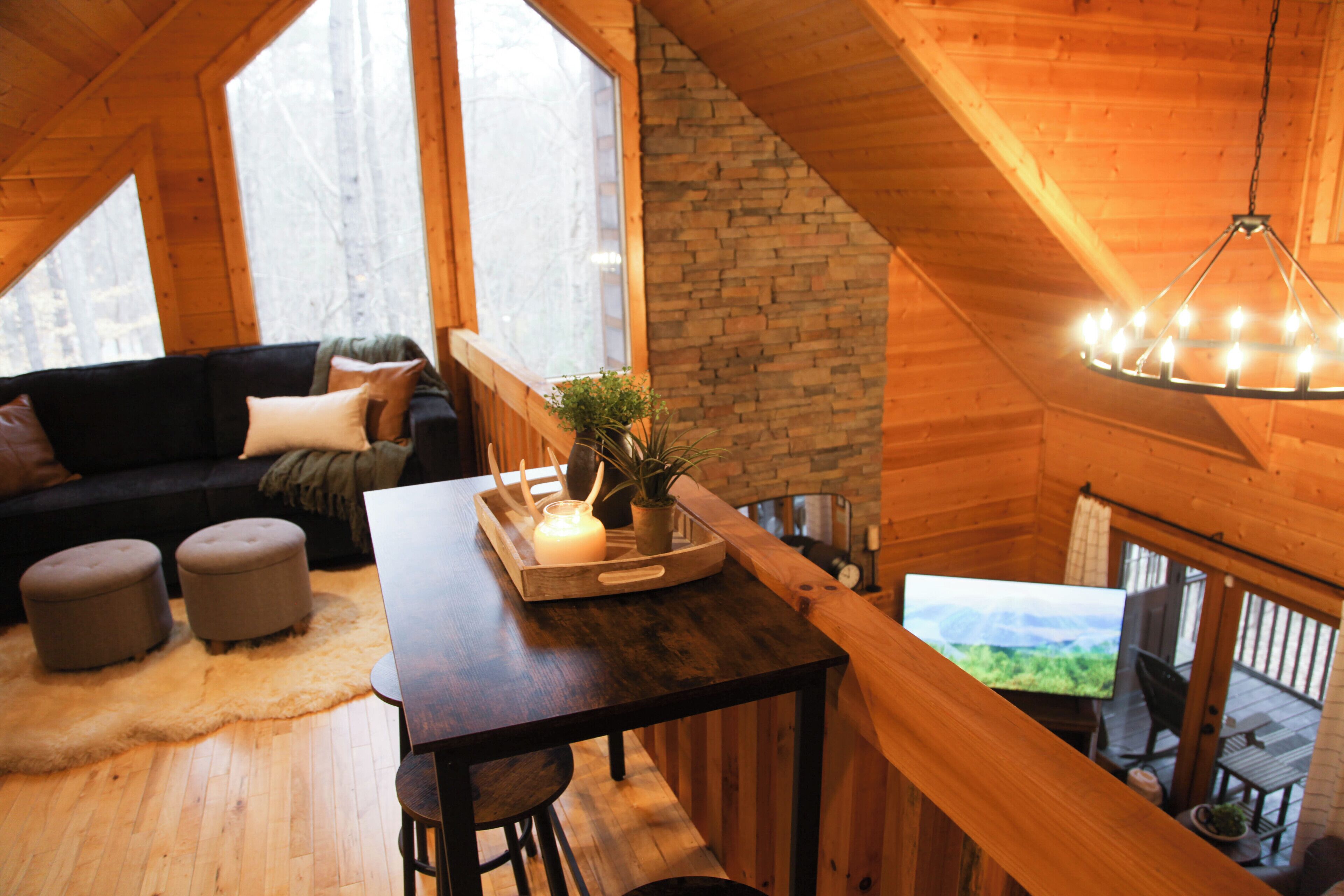 A cozy loft-style room with a wooden ceiling, a stone fireplace, a dark wooden table with a candle and a plant, a black leather sofa, and a view of the outdoors through large windows.