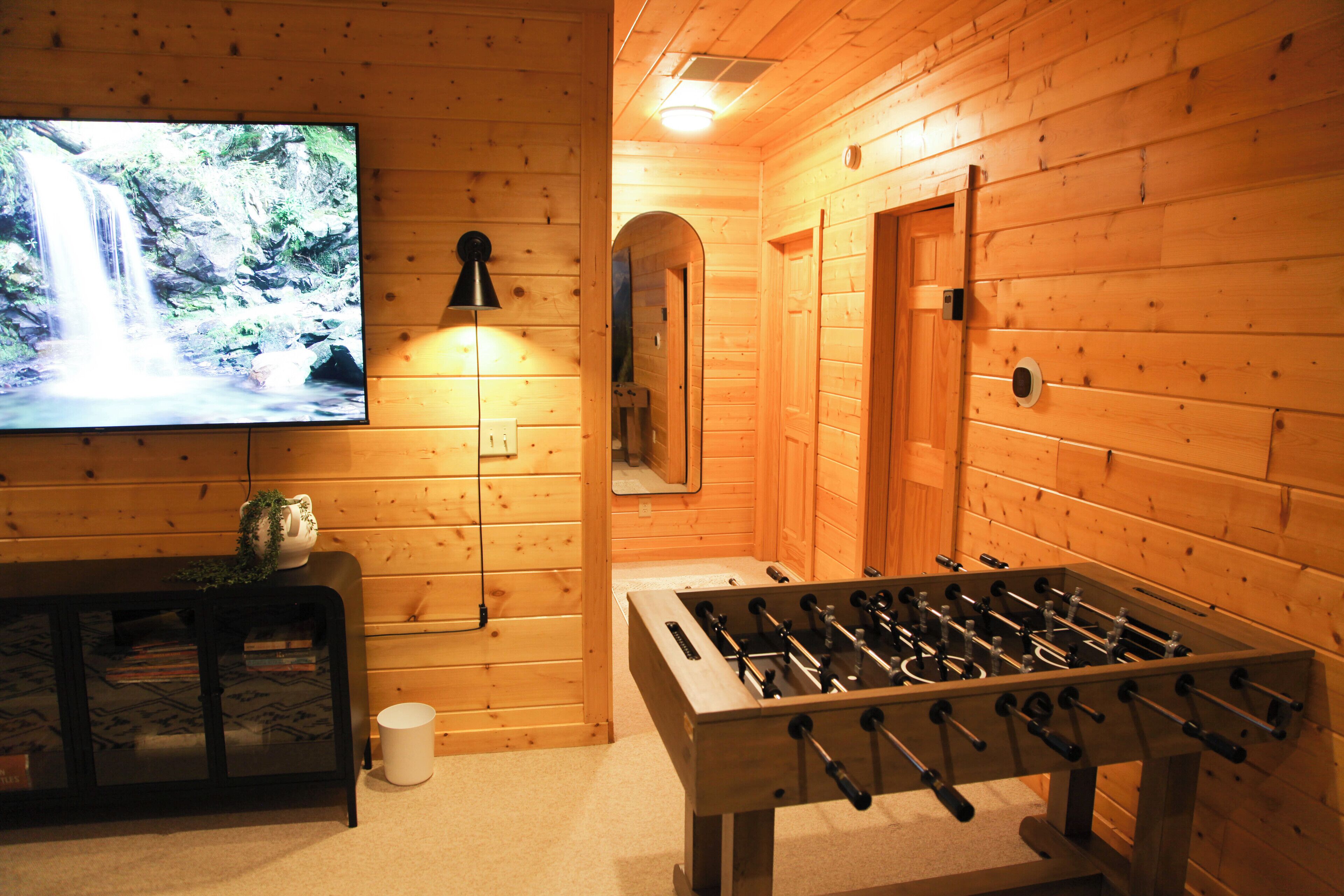 A wooden-floored room with a foosball table, a television displaying a waterfall, and a small table with a plant.