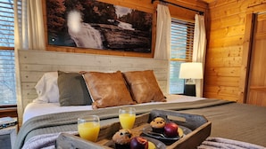 A cozy room with a wooden headboard, a bed with pillows, a tray with food and drinks, and a painting of a waterfall on the wall.