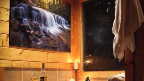 A wooden cabin bathroom with a large window, a painting of a waterfall, a bottle of wine, and a towel hanging on the wall.