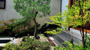 A traditional Japanese-style building with a wooden roof and a well-maintained garden featuring a pine tree, bamboo, and other greenery.