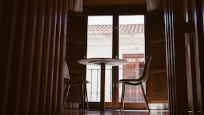 A room with a round table and two chairs, large windows with a view of a building with a tiled roof, and a hanging light fixture. - Los Mercaderes - Hotel & Spa (Talavera de la Reina)