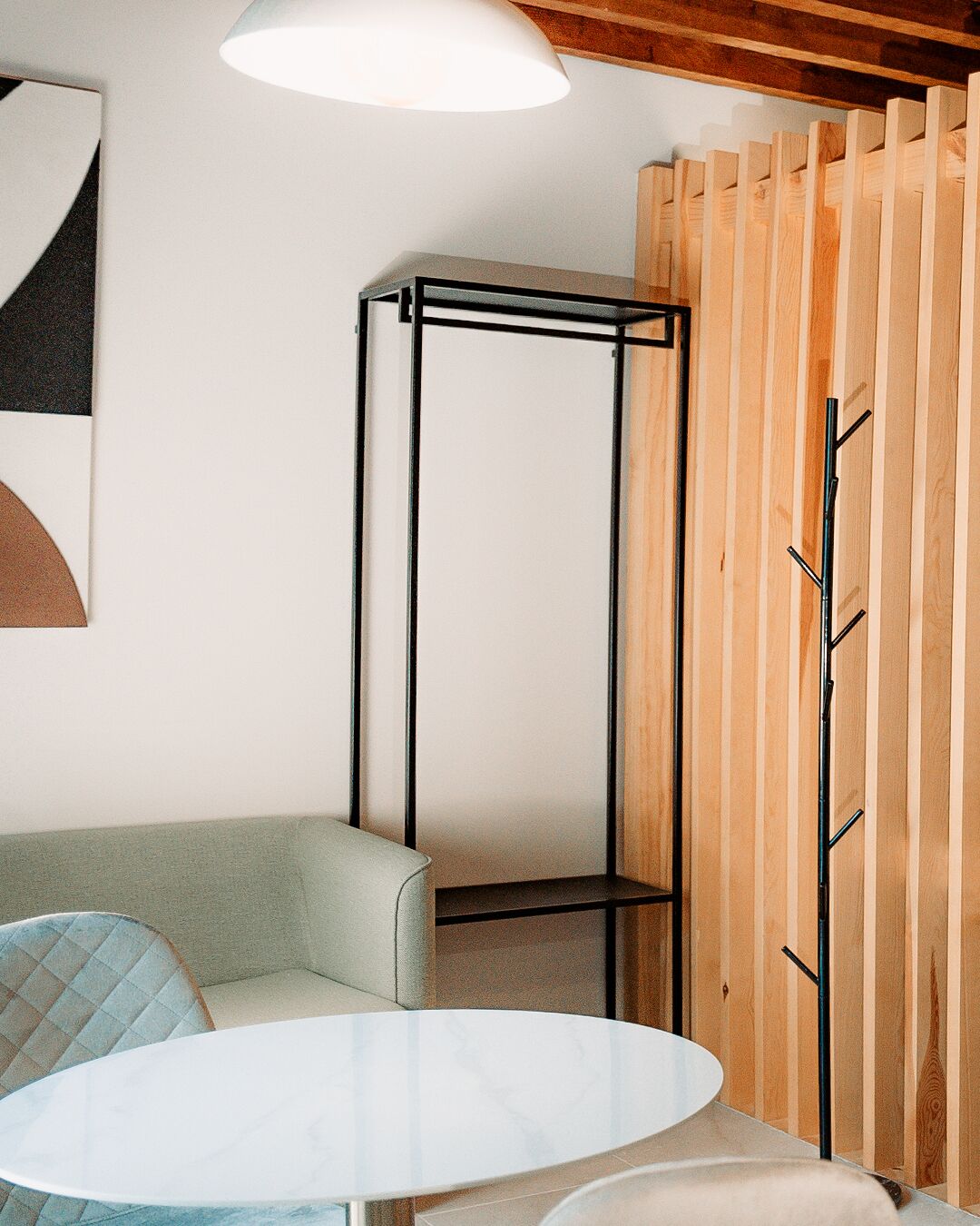 Una habitación de esquina con un sofá blanco, una pequeña mesa redonda, un soporte de metal negro y un separador de madera con listones.