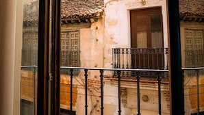 A view through a window of a stone building with a tiled roof, a wooden door, and a balcony with wrought iron railings. - Los Mercaderes - Hotel & Spa (Talavera de la Reina)
