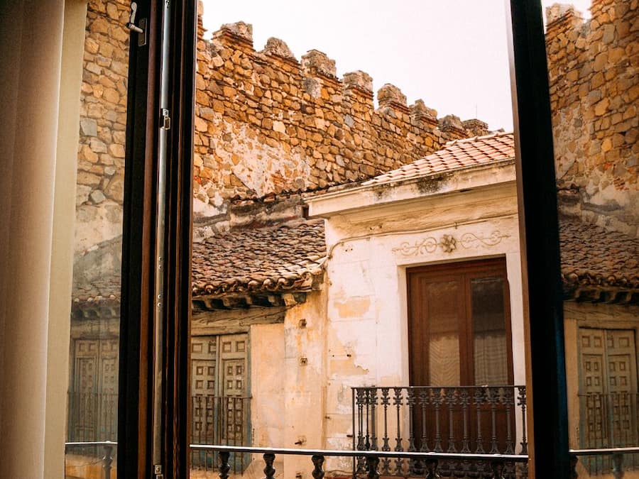 A view through a window of a stone building with a tiled roof, a wooden door, and a balcony with wrought iron railings.