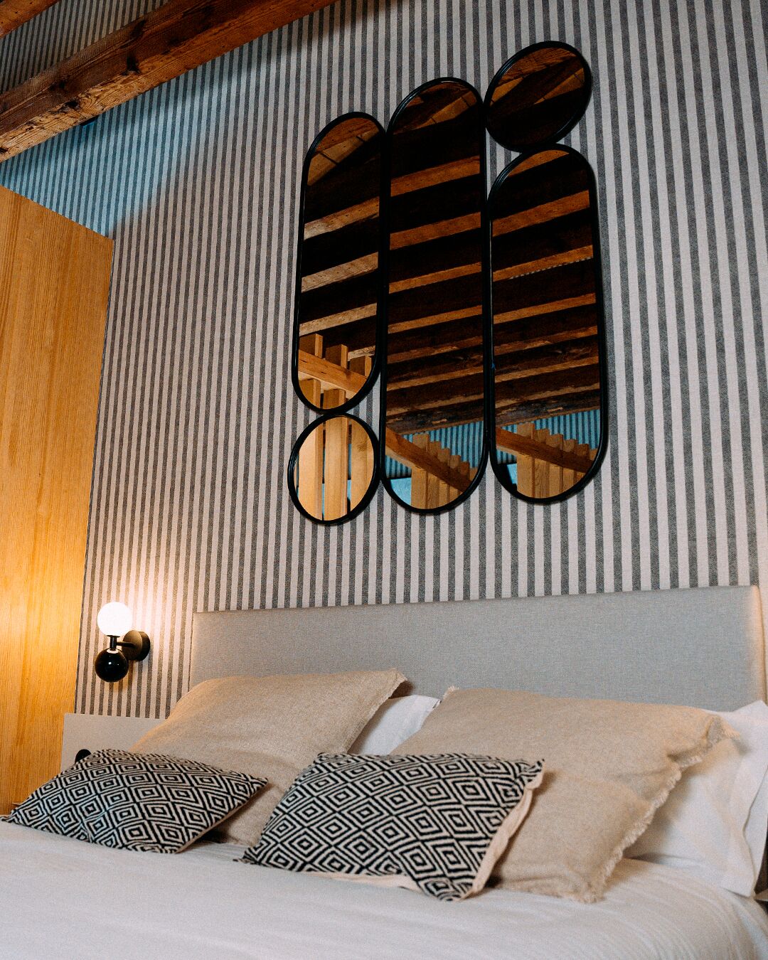 A hotel room with a striped wall, a bed with patterned pillows, and a wooden ceiling beam.
