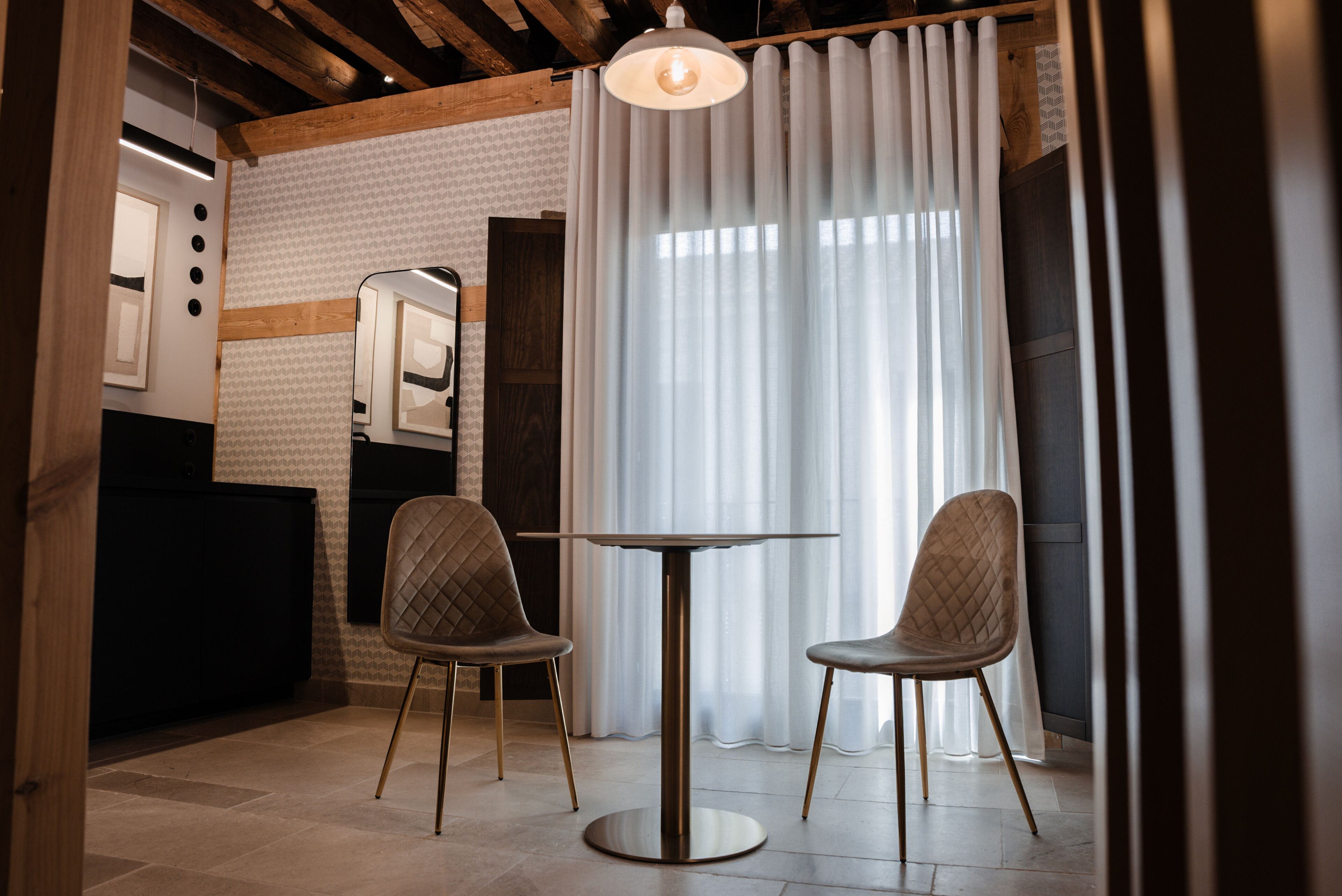 A modern interior with a round table and two chairs, a wooden ceiling, and a large window with sheer curtains.