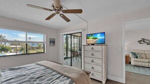 Un dormitorio con cama, televisor, ventilador de techo, puerta corrediza de vidrio y vista a una piscina y palmeras.