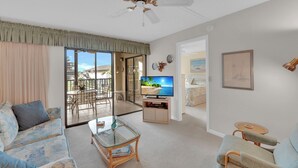 A spacious living room with a large TV, a balcony with a view, and a ceiling fan. - SUMMER/FALL DISCOUNT!!!  2B/2B Fairview Isles 123 Condo, Walk to Beach, Heated Pool! (Fort Myers Beach)