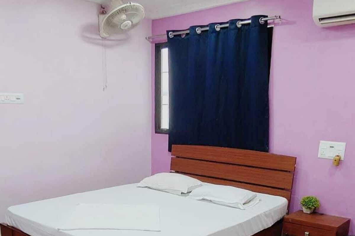 A bedroom with a single bed, a ceiling fan, an air conditioner, a window with a dark curtain, and a small bedside table with a plant.