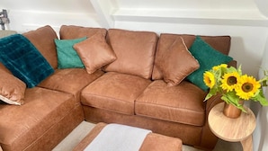 A brown sectional sofa with teal and blue cushions, a small round wooden side table with a vase of sunflowers, and a chevron-patterned rug. - Central Tenby 3 Bed Apt Sleeps 6 to 8 Dog Friendly (Tenby)