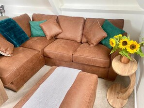 A brown sectional sofa with teal and blue cushions, a small round wooden side table with a vase of sunflowers, and a chevron-patterned rug. - Central Tenby 3 Bed Apt Sleeps 6 to 8 Dog Friendly (Tenby)