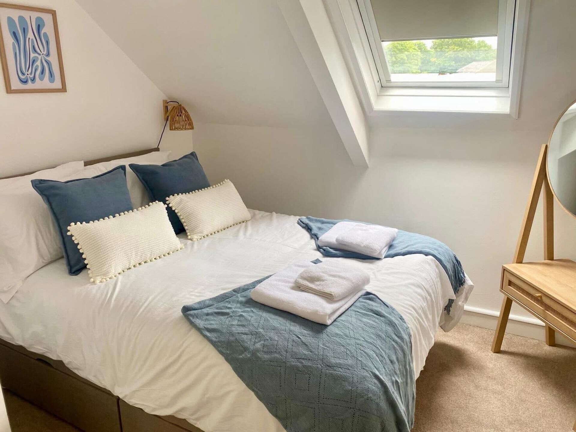 Ein ordentlich bezogenes Bett mit Kissen und Decke, ein kleiner Tisch mit Stuhl und ein Fenster mit Blick auf Grünflächen.