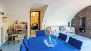 A modern living room with a blue sofa, a glass coffee table, and a bookshelf. There is a dining area with a blue table and chairs. The room has a curved ceiling and a stone wall.