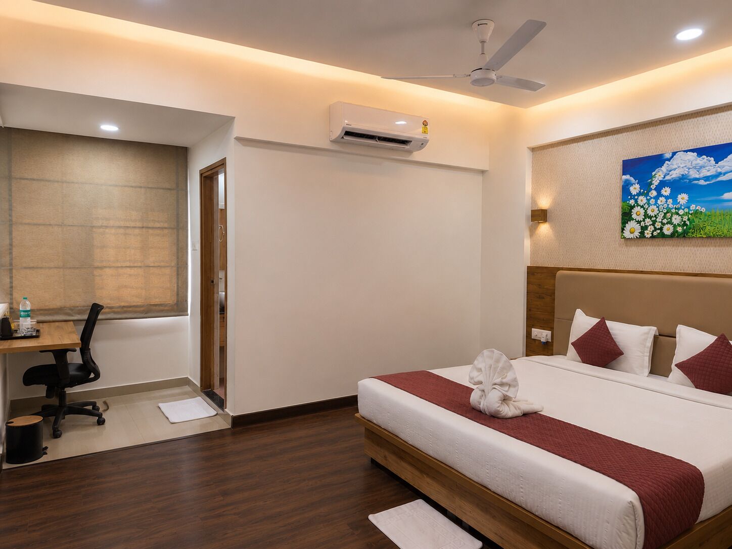 A hotel room with a bed, a desk, a chair, an air conditioning unit, and a painting on the wall.