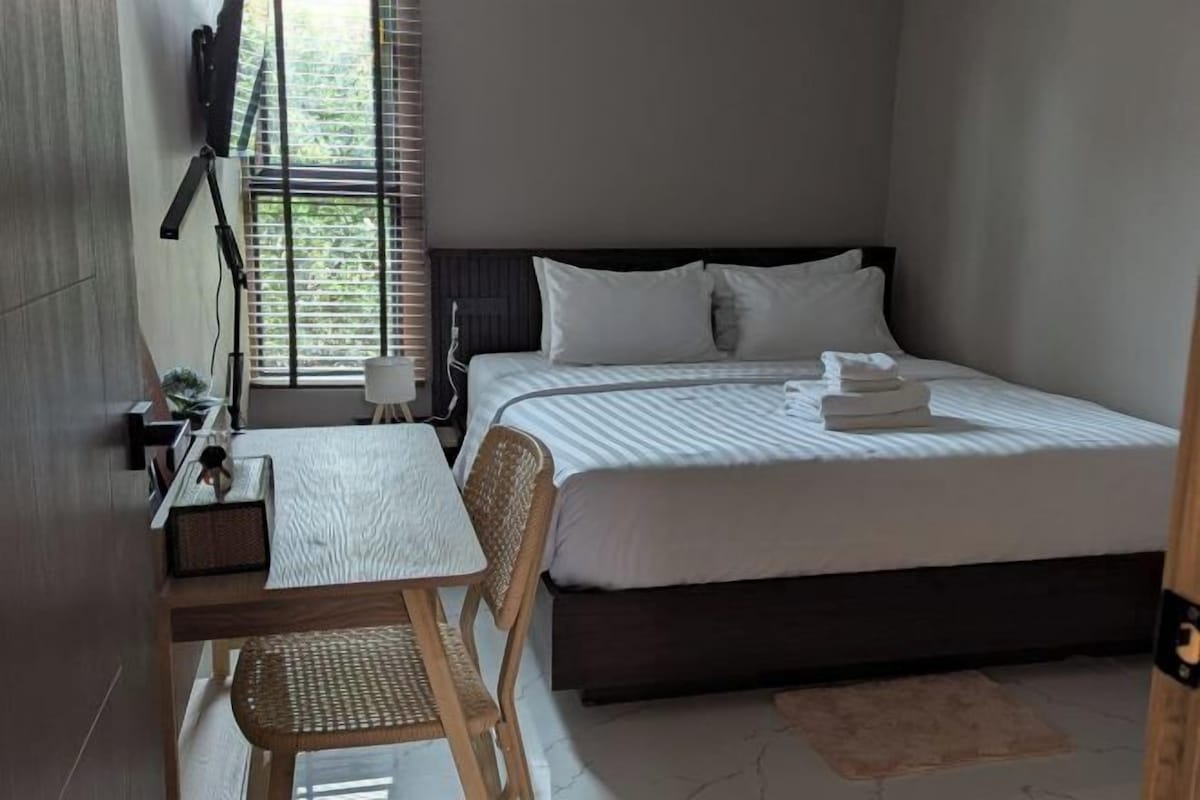 A hotel room with a bed, a small table with a chair, a TV mounted on the wall, and a window with blinds.